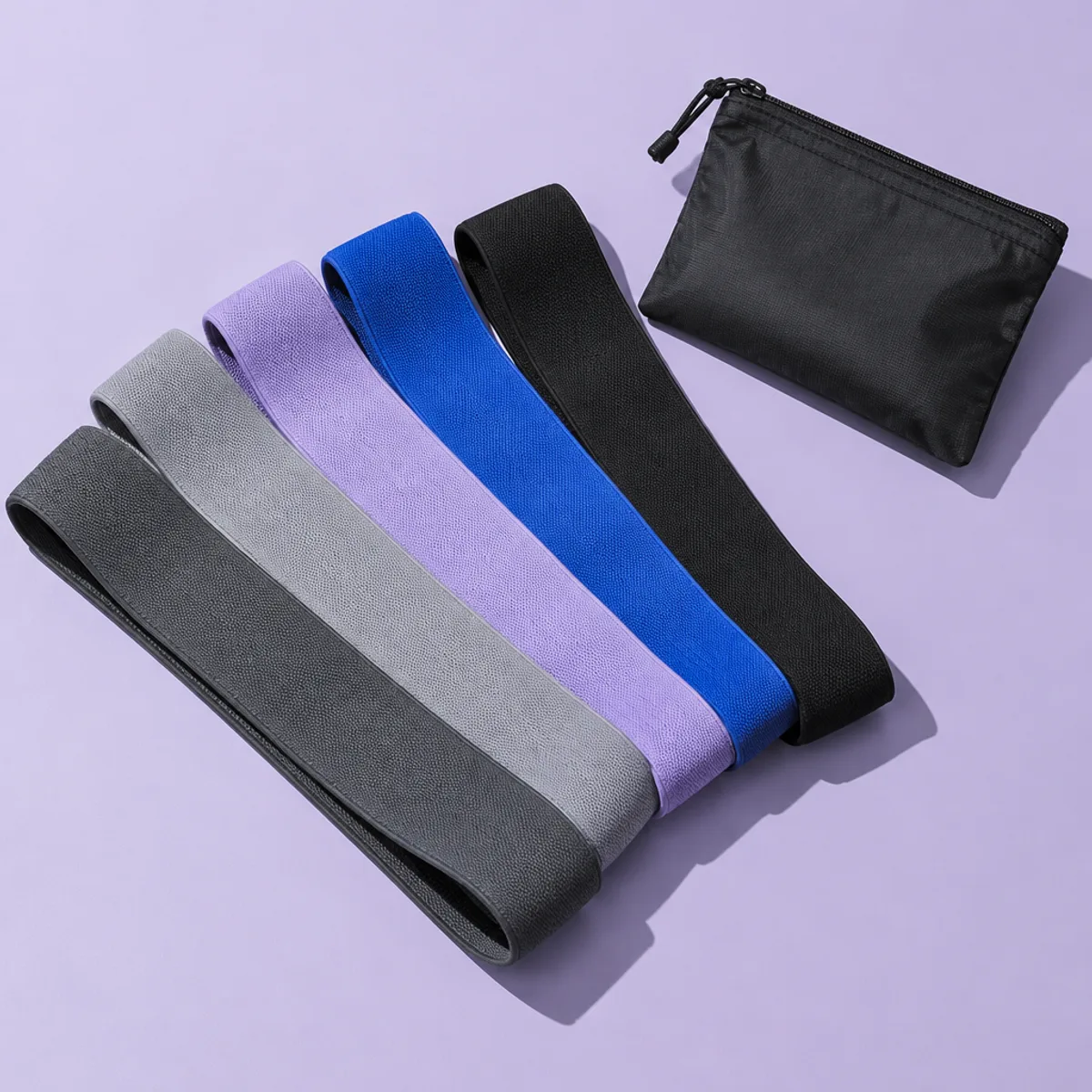 Five fabric loop resistance bands in charcoal, grey, lilac, blue and black beside a small black pouch on a lilac background